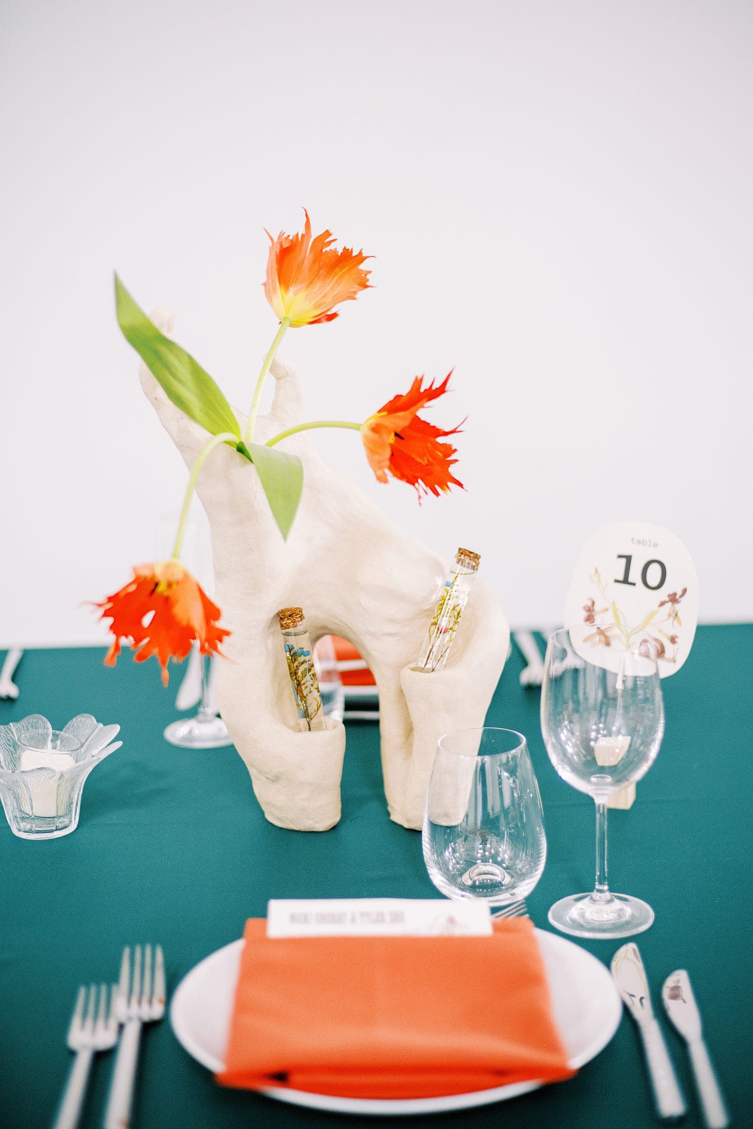 Styled wedding reception place setting with orange parrot tulips in a sculptural white vase and a folded orange napkin on a teal linen tablecloth, photo by Kelly Prizel