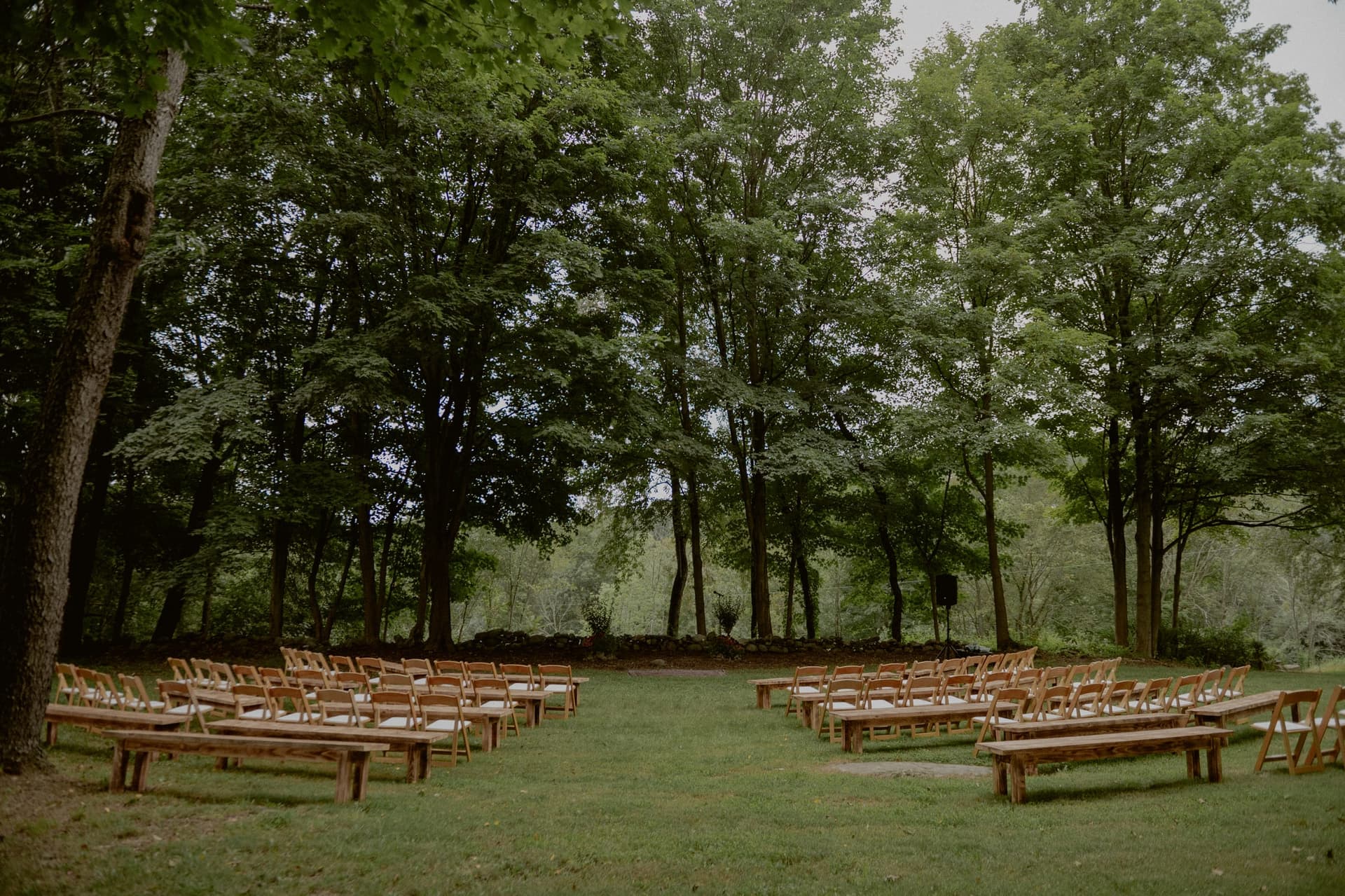 Outdoor wedding ceremony with wooden benches and chairs arranged in a semicircle on a grass lawn surrounded by mature trees, photo by Chellise Michael Photography